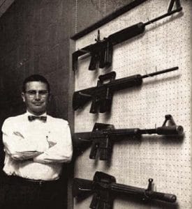 Eugene stoner standing next to a wall of ar15 rifles
